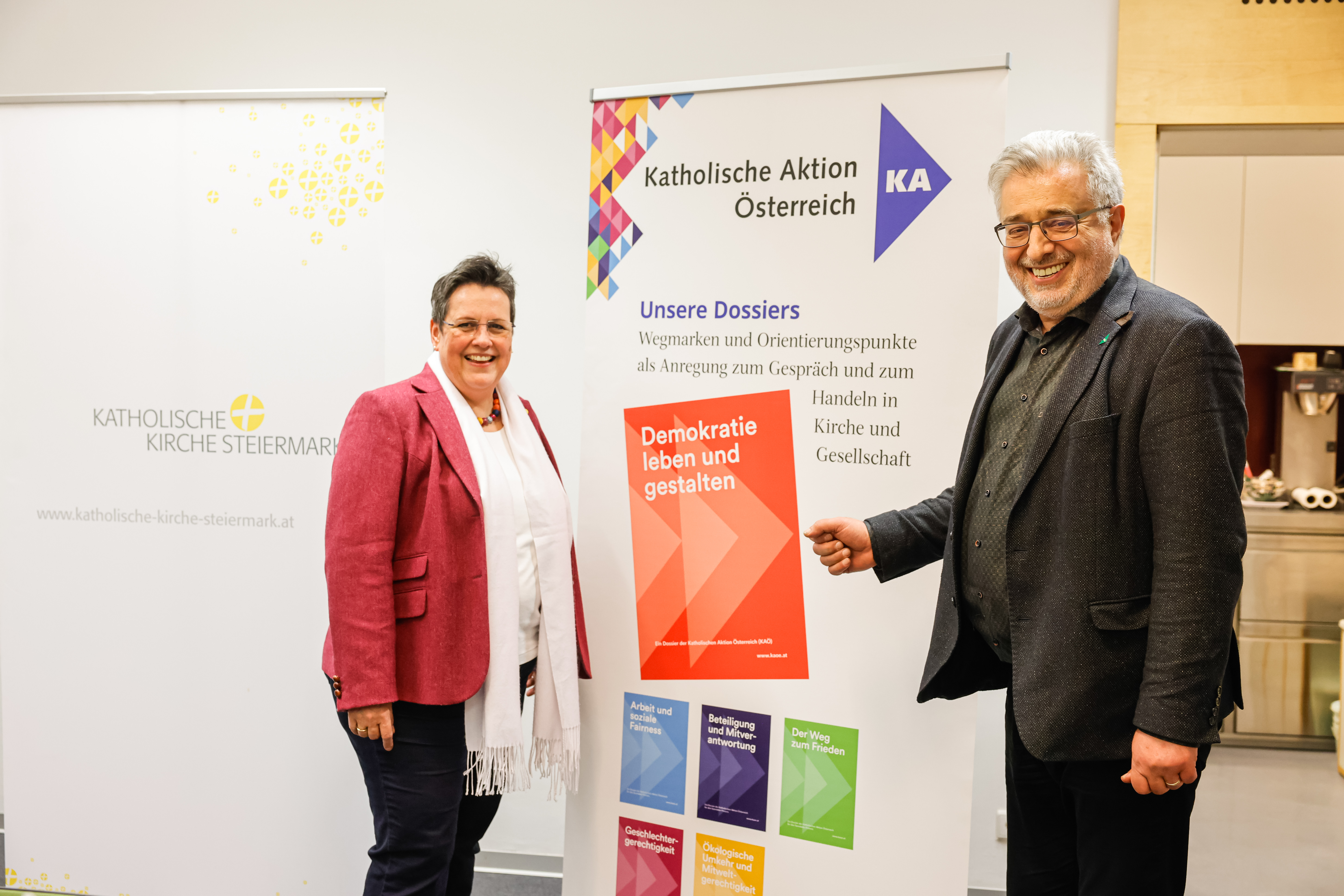 Anna Hollwöger und Andreas Gjecaj stehen vor einem Rollup, auf dem die Dossiers der KAÖ abgebildet sind